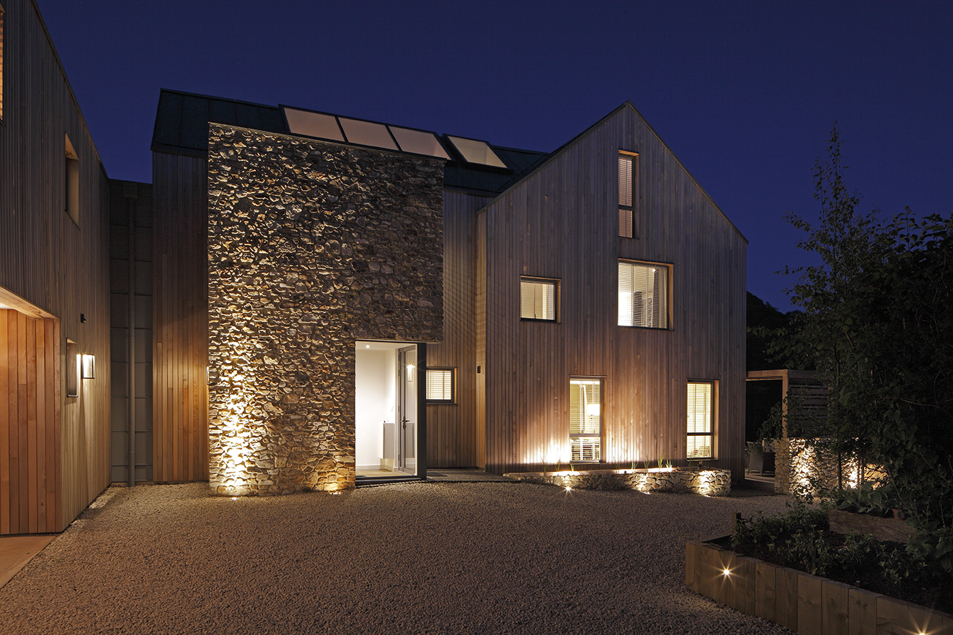 Modern house in timber and stone, at dusk with lights in the windows and exterior lighting against a dark blue sky.