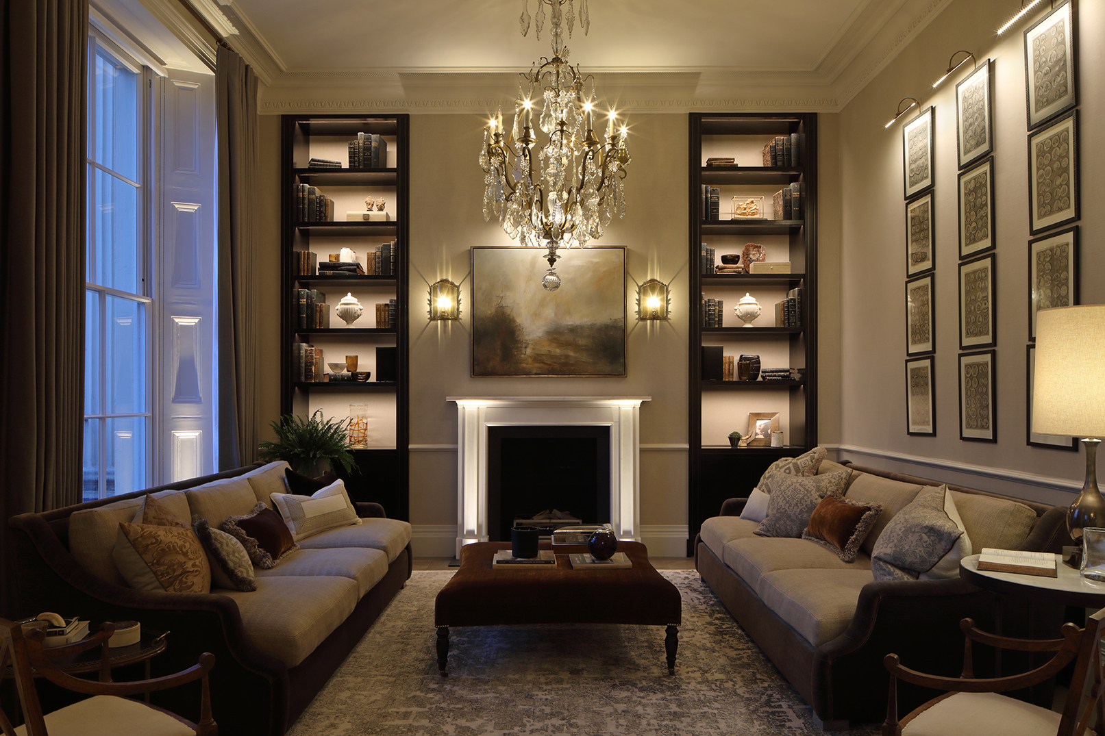 Grand regents park drawing room with two sofas either side of marble fireplace and chandelier. Recessed lit shelving along back wall. Blue dusk light at the full height sash window on the left.