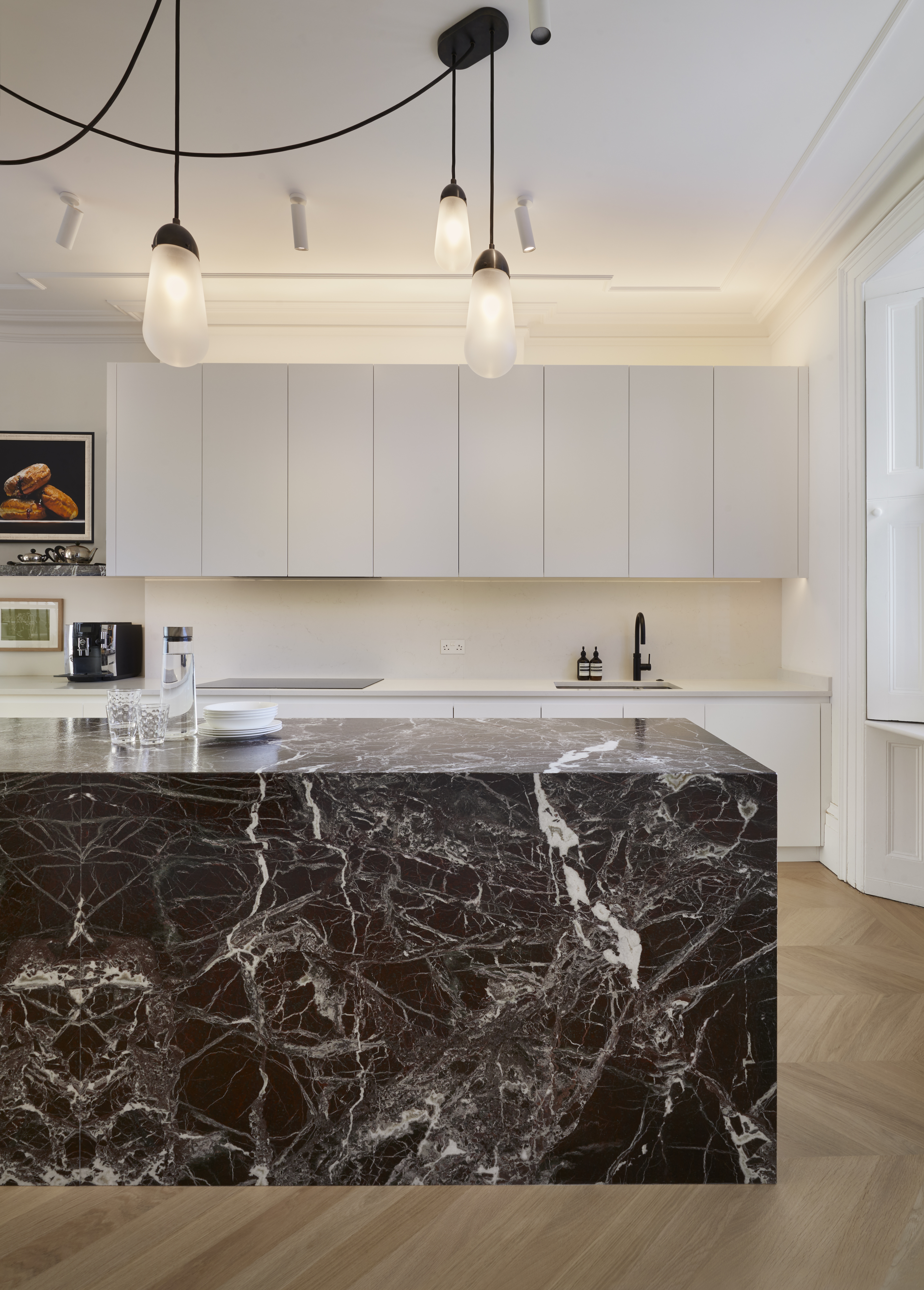 The rear view of a marble-clad kitchen island with white painted wall cabinets above the sink area against the wall in the background