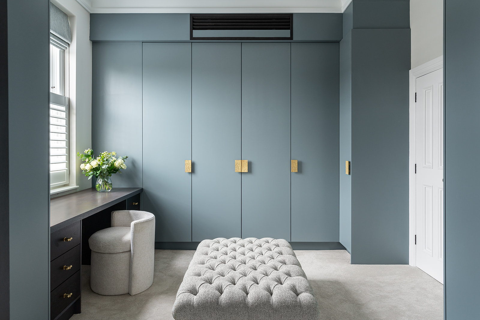 Elegant Walk-In Wardrobe Design in the principal suite of a Barnes Family Home - Luxurious walk-in wardrobe with custom blue cabinetry, handcrafted bespoke gold textured handles, and a plush tufted ottoman using fabric from Chase Erwin. Designed by an interior design studio in Barnes, London.