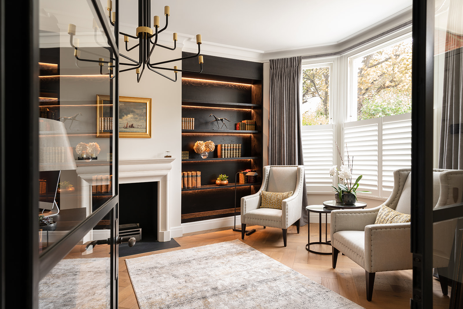 Sophisticated home office and library with bespoke joinery and ambient lighting – A refined study space in a prestigious Barnes home, featuring custom bookshelves, a statement Andrew Martin chandelier, and plush Wing-back seating for a timeless, elegant atmosphere.