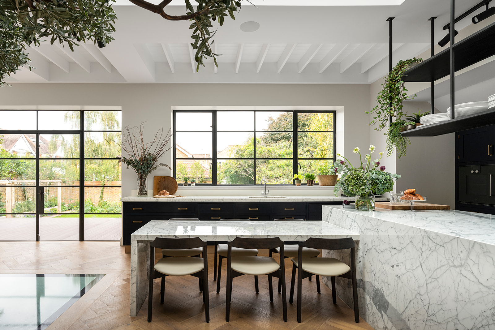 Luxury kitchen with marble island and black cabinetry in Barnes – A bespoke kitchen designed for a grand family home in Barnes, featuring an elegant marble island, dark cabinetry with brass handles, and large Crittall-style windows opening to a lush garden.