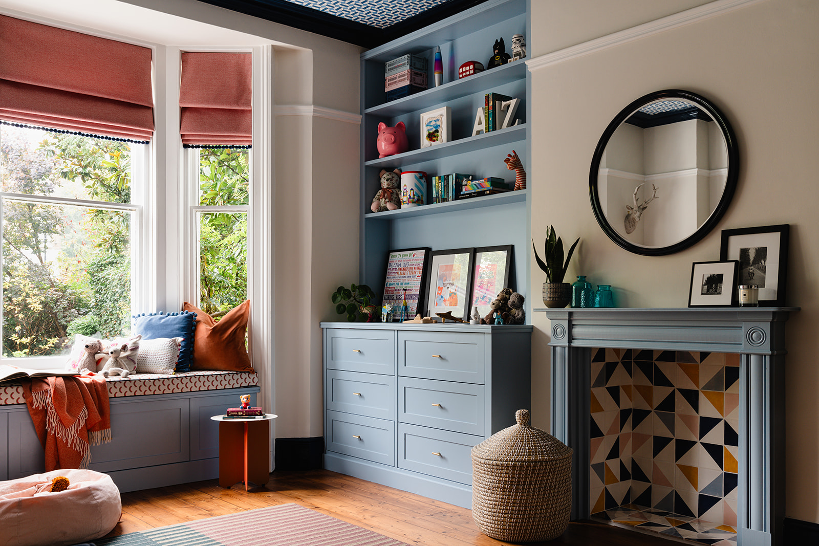 Beautifully designed kids' playroom with custom-built blue cabinetry, a bold patterned ceiling, and cosy bay window seat. Warm terracotta blinds and soft textiles add depth and comfort.