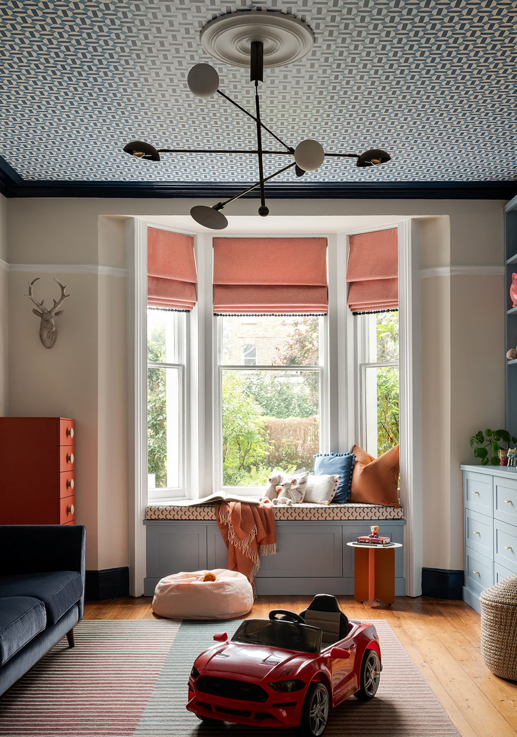 Stylish and playful children's playroom with a cosy window seat, bold patterned ceiling, and pale blue built-in storage. Features warm terracotta accents, soft textiles, and a modern ceiling pendant for a fun yet sophisticated space.