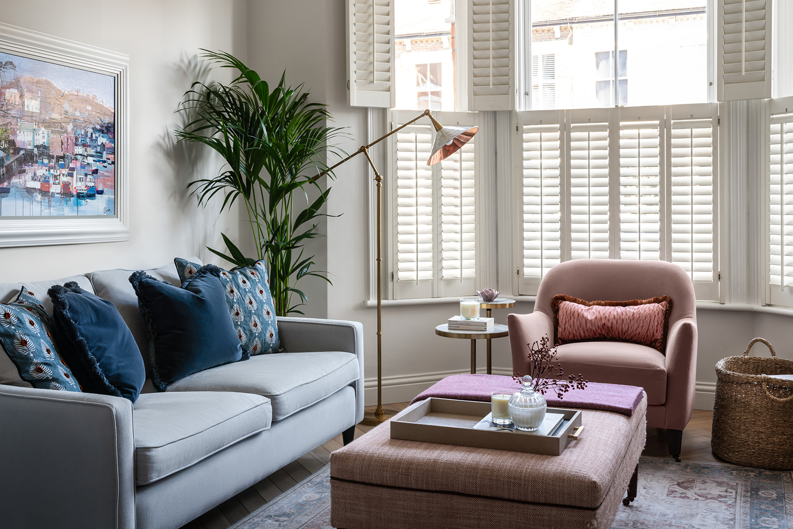 Bright and airy living room featuring a soft blue-grey sofa, plush blue and pink accents, and large bay windows with plantation shutters.