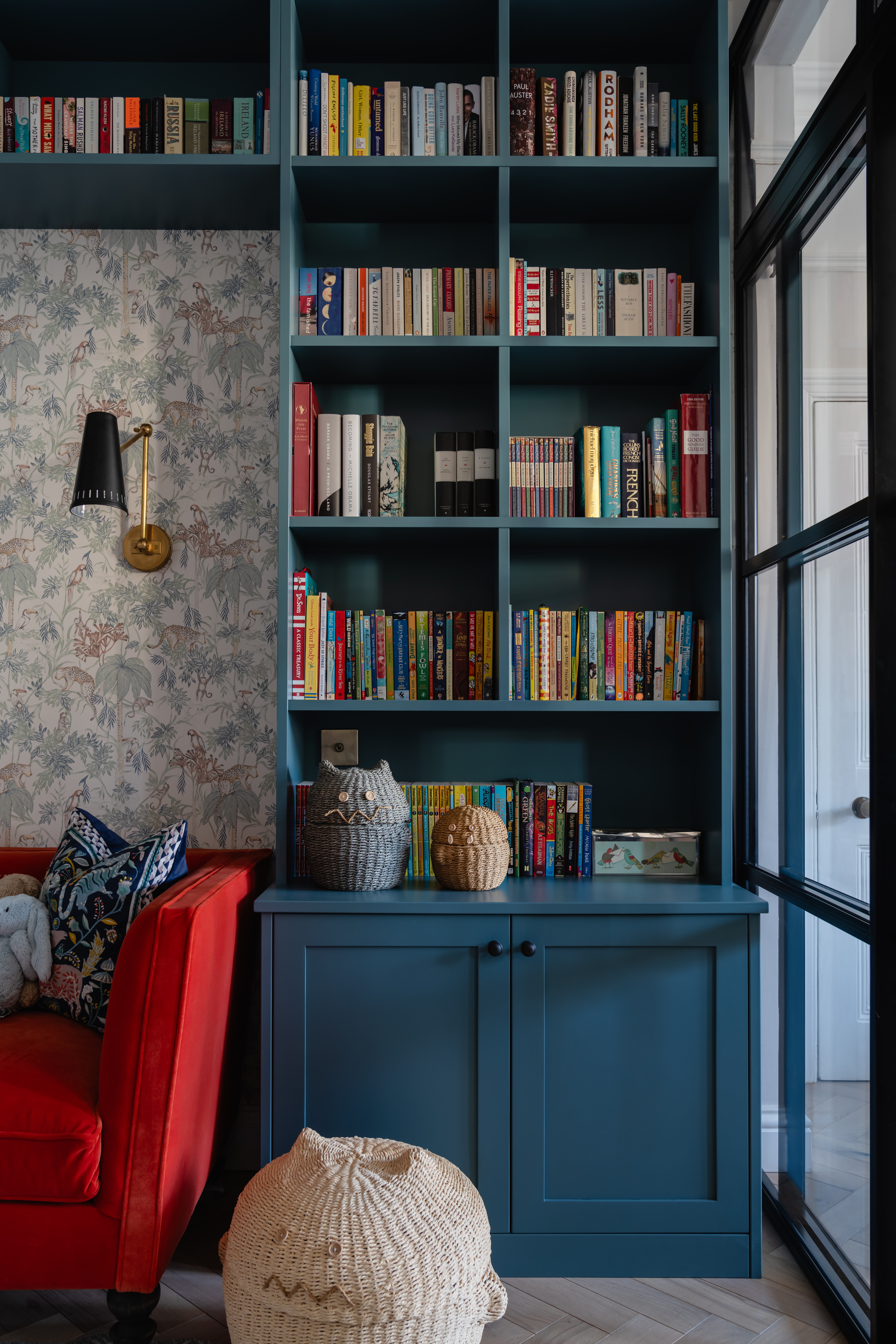 Charming playroom corner featuring custom blue joinery painted in Etruria (Little Greene), a cosy reading nook, and playful accessories, complemented by a jungle-inspired wallpaper and an elegant brass wall light (Andrew Martin "Antonio" Wallight), adding warmth and character to the space.