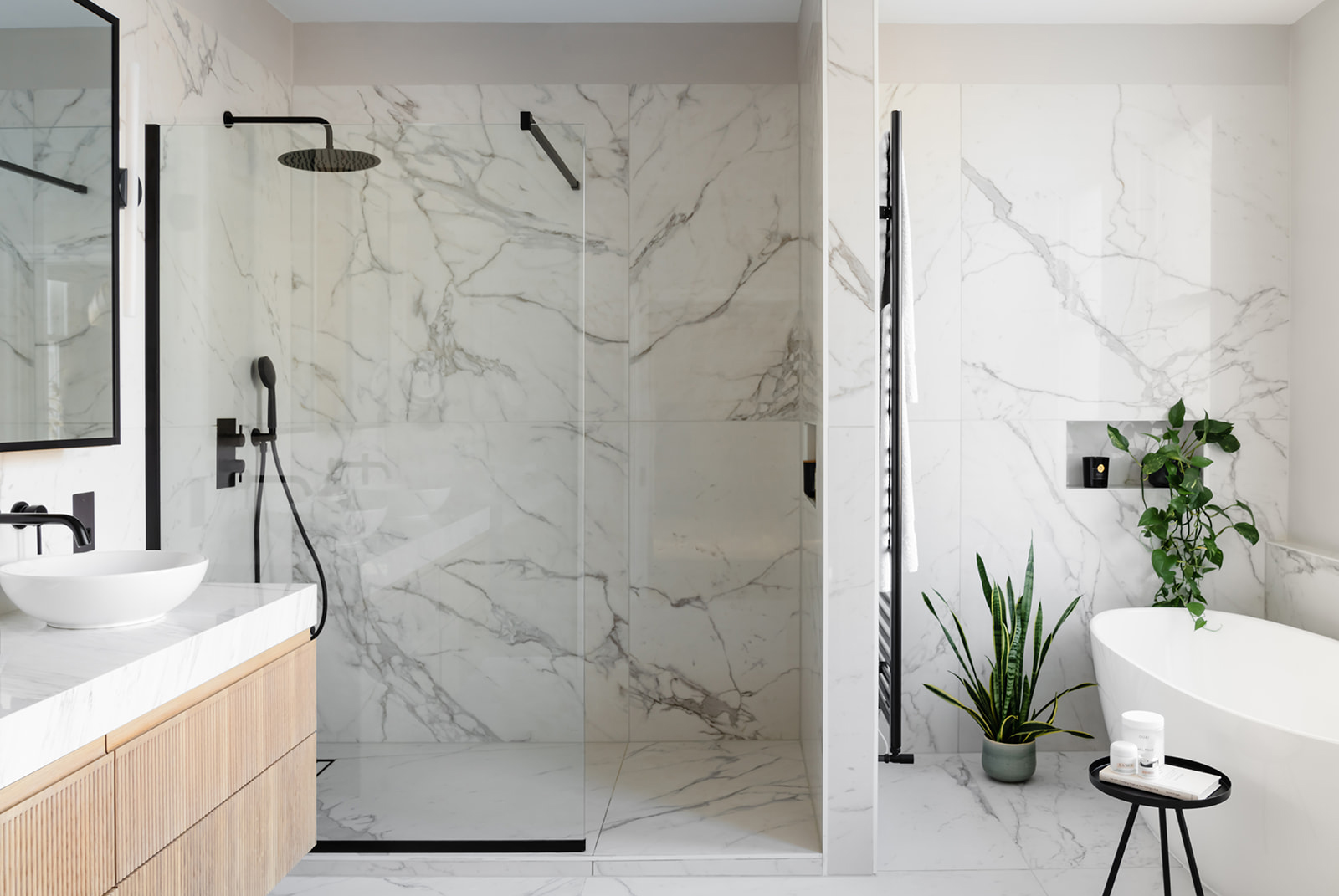 A beautifully designed bathroom with a large walk-in shower, a freestanding bath, and verdant plants. Cararra marble walls and black fixtures provide a timeless and elegant aesthetic.