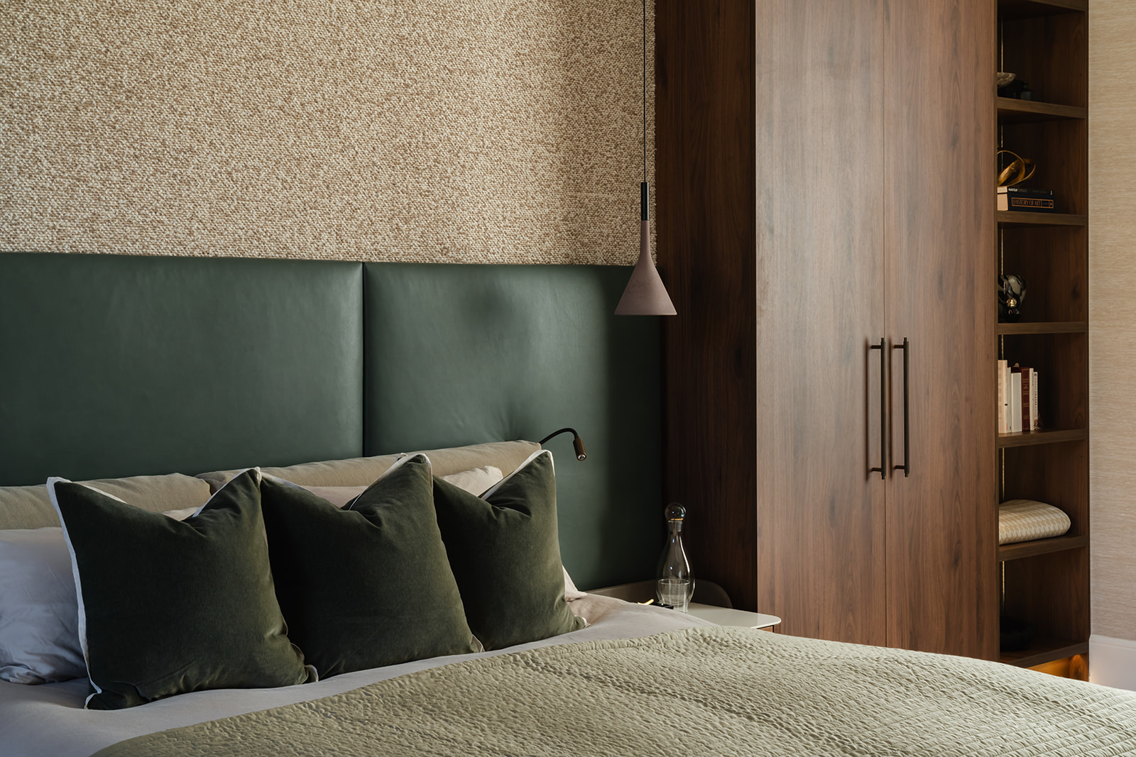 A beautifully designed bedroom featuring a green leather headboard, bespoke built-in wardrobes in walnut with open shelving, and grasscloth textured wallpaper in ivory. Floor-length curtains frame a large window, softening the natural light for a calming retreat.