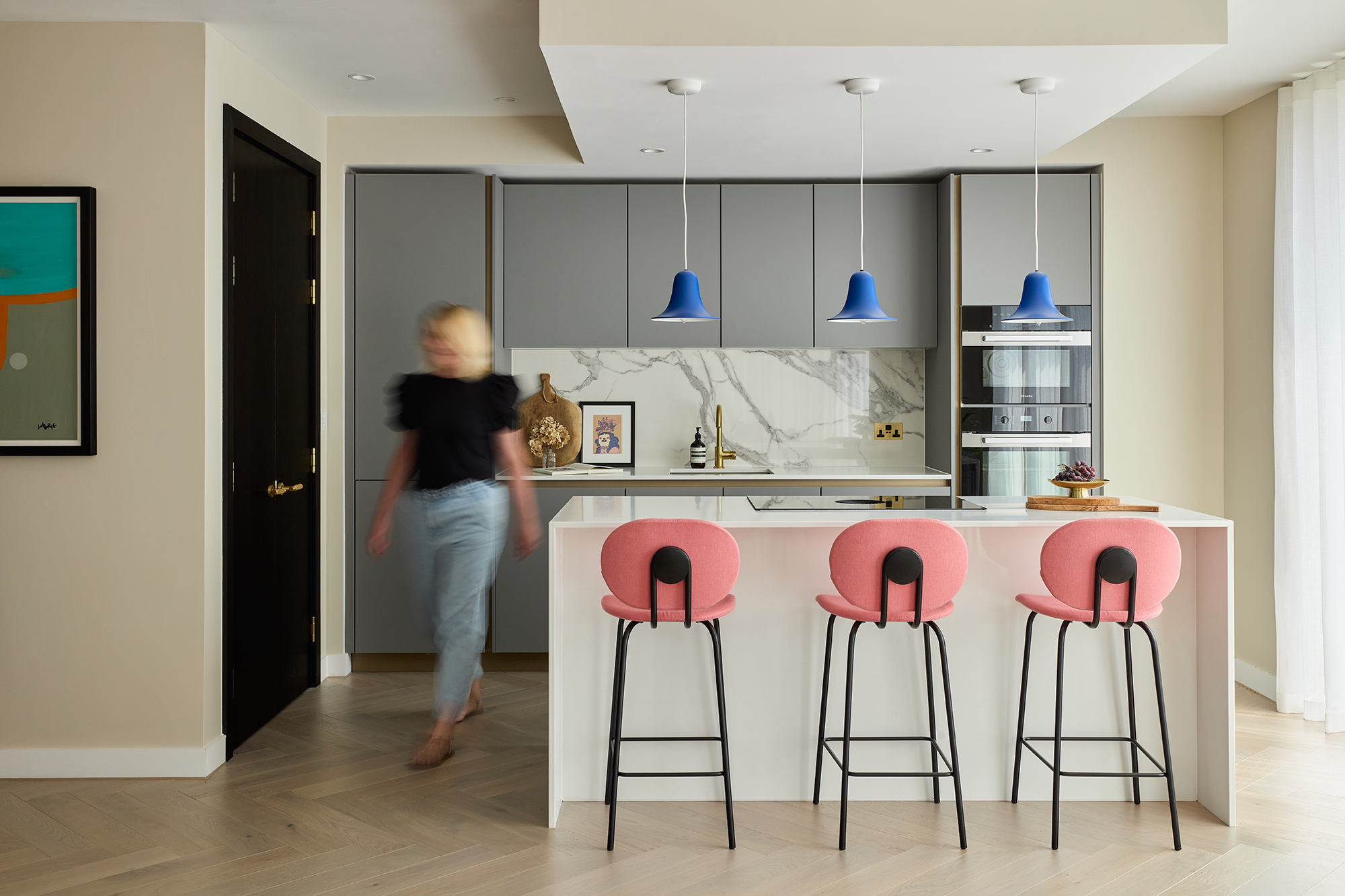 Stylish kitchen island in a Triptych Bankside apartment, featuring pink bar stools, a white quartz worktop, and blue pendant lights for a vibrant touch.