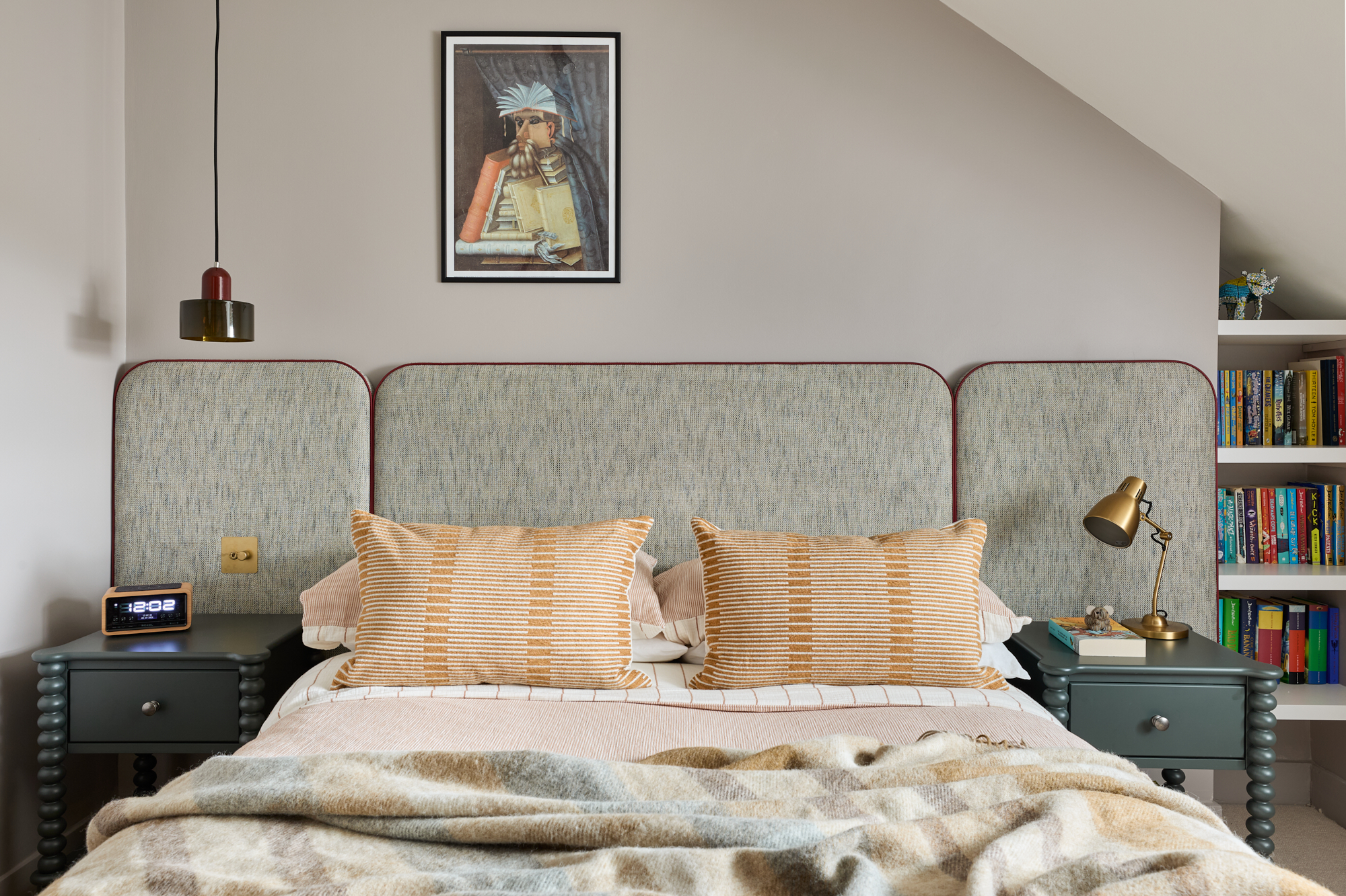 A Stylish teenage bedroom featuring a three-panel upholstered headboard in soft blue-grey fabric with contrasting terracotta piping. The bed is styled with mustard and cream striped cushions, layered terracotta linen bedding, and a cosy wool throw in neutral and earthy shades. Framed artwork above the bed adds a personal and artistic touch.