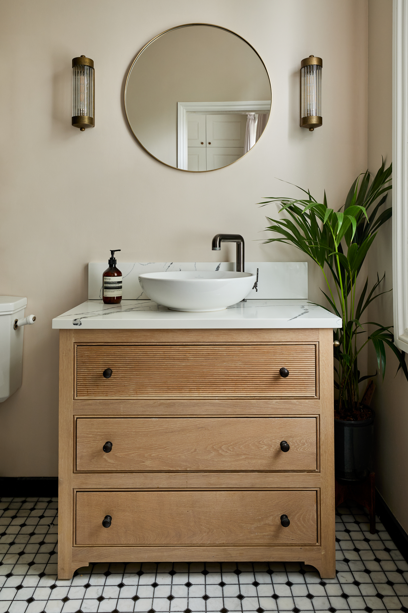 A beautifully styled bathroom vanity that exudes sophistication. The oak cabinet with fluted drawer fronts is topped with a pristine white-black veined marble countertop, supporting a sleek vessel sink and bronze tap. Above the vanity, a large round brass framed mirror reflects the room’s warm tones, while elegant brass wall sconces add ambient lighting. A lush green plant in the corner introduces a fresh, organic touch to the space. Calacatta marble Victorian octagon mosaic floor tiles reflect the property