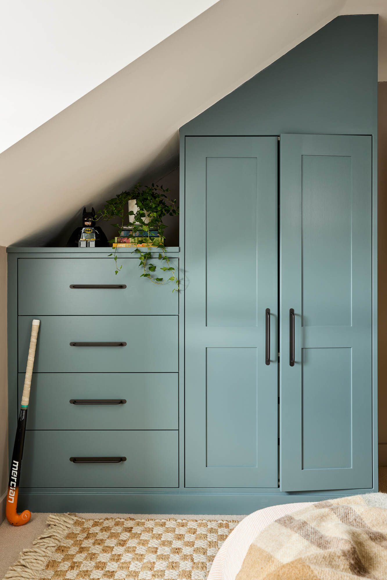 A bespoke, built-in wardrobe and chest of drawers in Farrow & Ball’s De Nimes, designed to fit seamlessly into the attic’s sloped ceilings. Sleek Corston bronze handles add a modern touch, while open shelving provides a display space for books, plants, and personal decor. A classic Batman figurine adds a playful element to the otherwise sophisticated design.