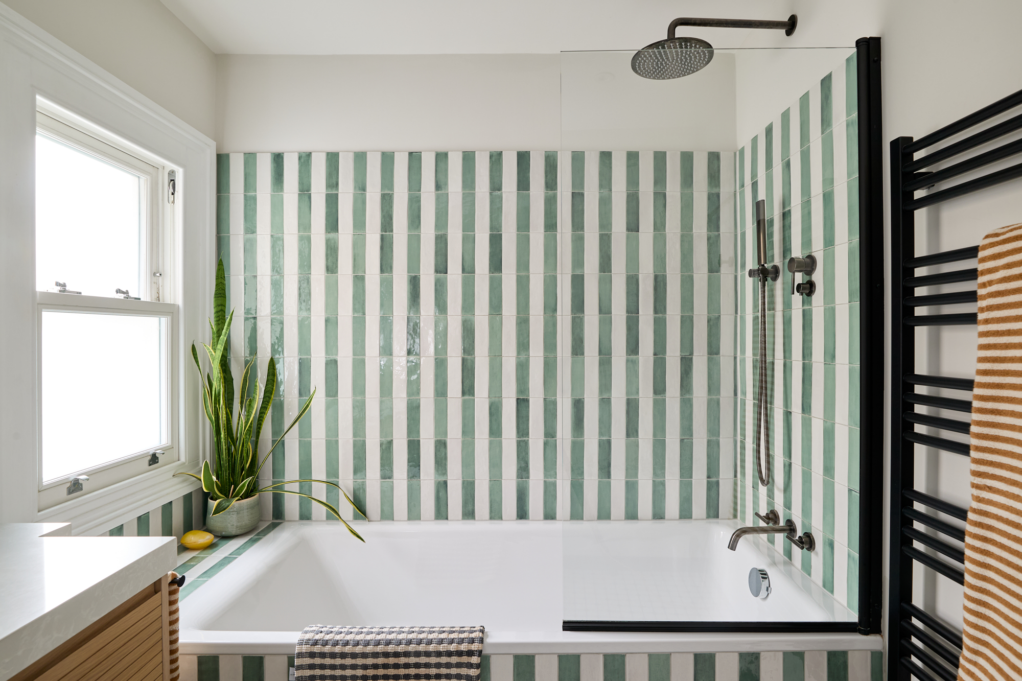 A stylish and modern shower over a bath featuring striking green-and-white vertically striped tiles that add a playful yet refined element to the space. A sleek black-framed glass partition keeps the design airy and open, while the rainfall showerhead and minimalist gunmetal fixtures provide a luxurious experience. A black heated towel rail with neatly folded ochre striped towels adds warmth and functionality to the contemporary bathroom.