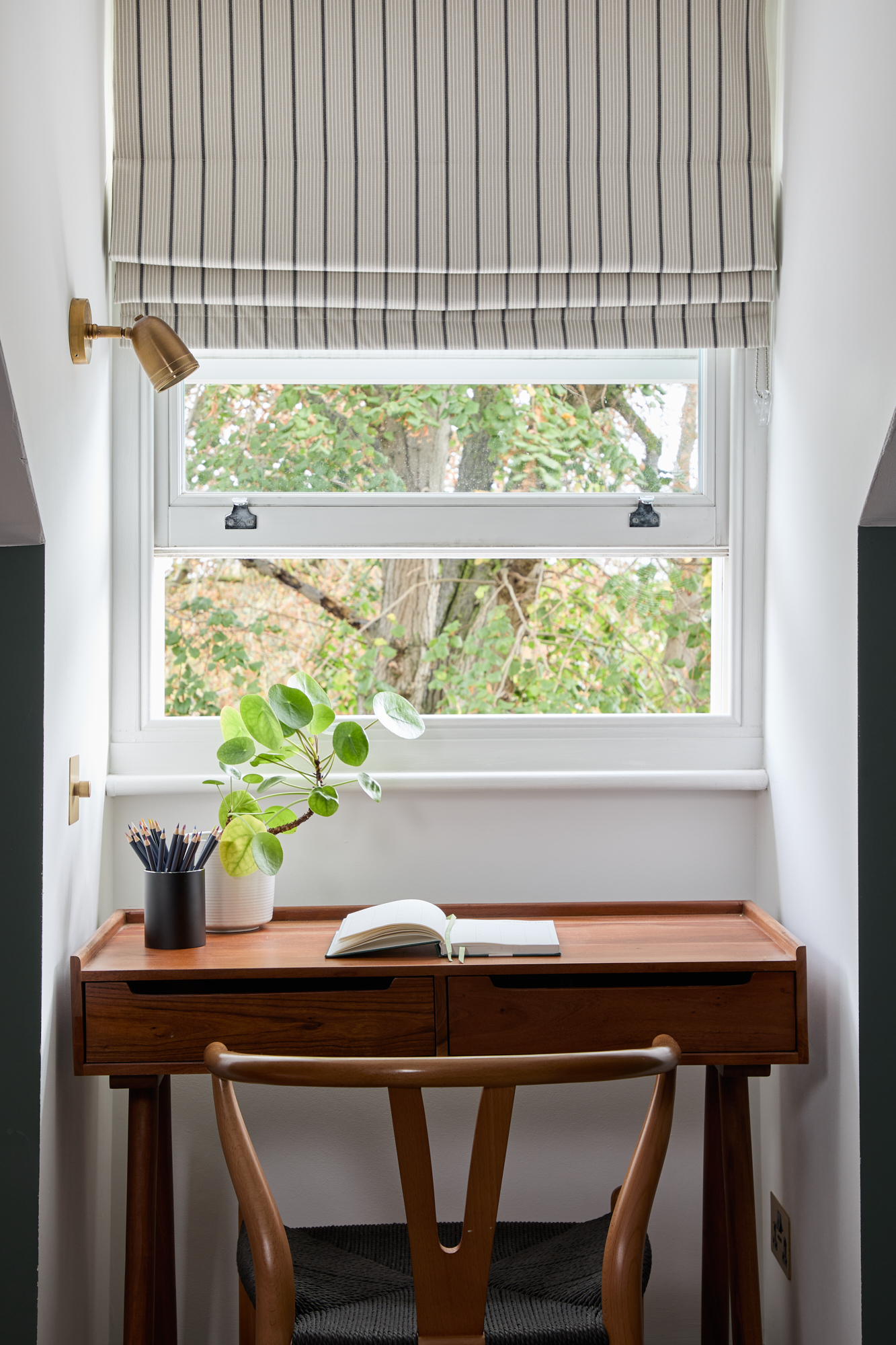 A beautifully crafted wooden desk sits beneath a bright window, creating an inviting workspace. The desk is styled with an open notebook, a sleek pencil holder, and a lush green plant, adding a fresh and natural touch. A classic wooden chair with a woven seat complements the space, offering both comfort and timeless design. Soft natural light enhances the cosy yet functional aesthetic of this home office nook.