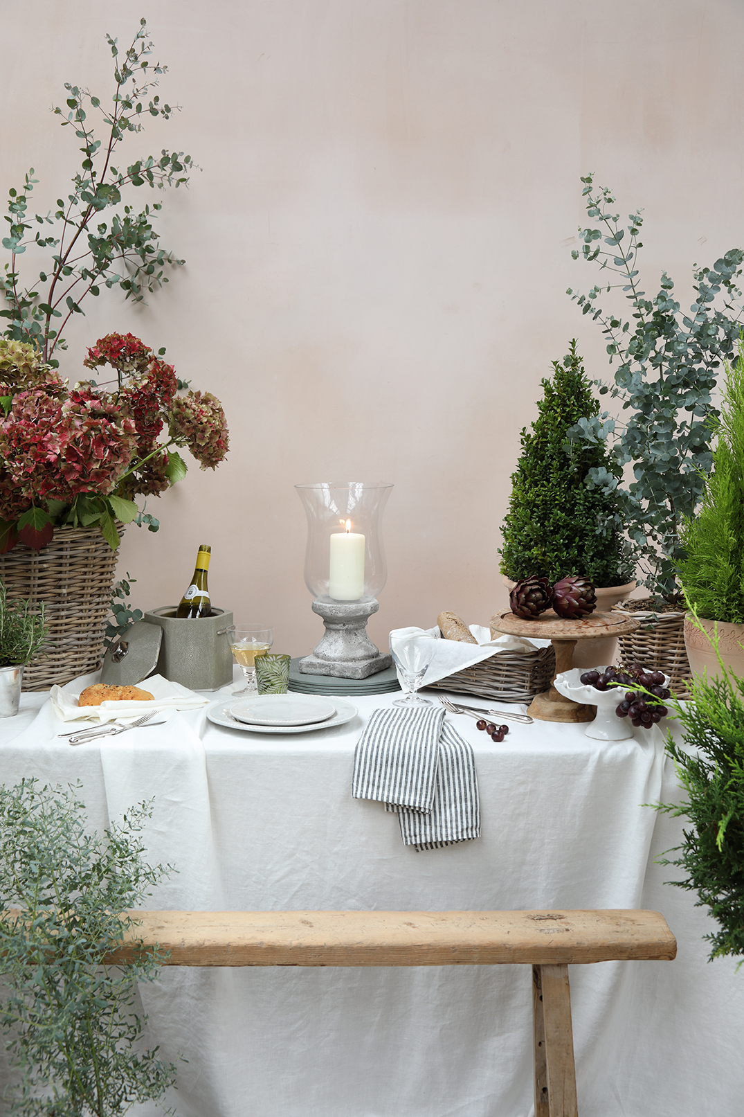 Still life table setting on a white linen tablecloth with a bench in front  surrounded by plants and flowers.