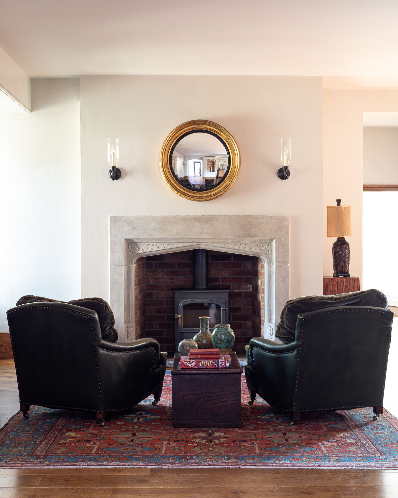 Elizabethan stone fireplace with wall lights, round mirror and leather club chairs in hallway.