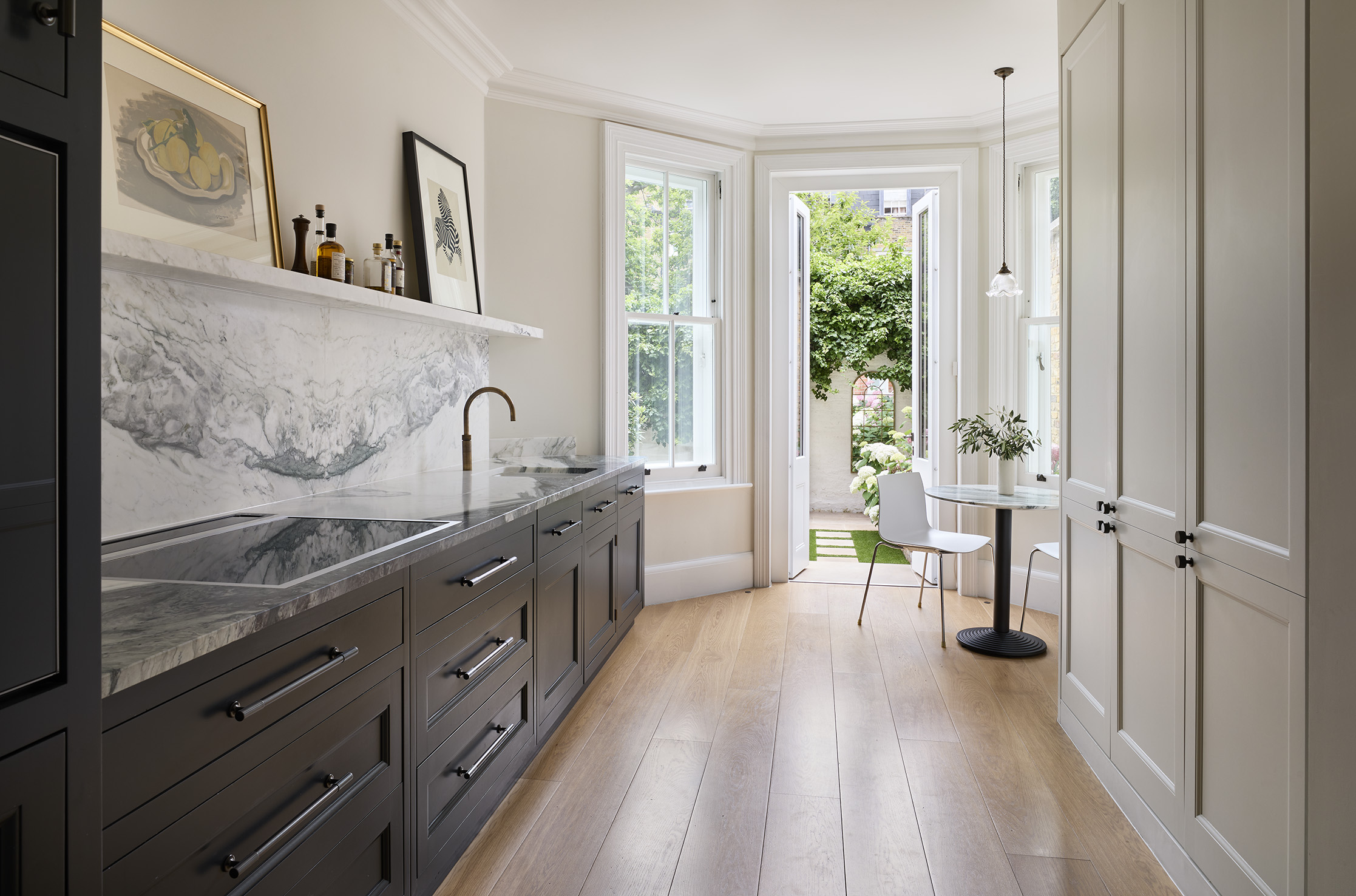 A view through a galley kitchen with dark grey & white cabinets to French doors at one end