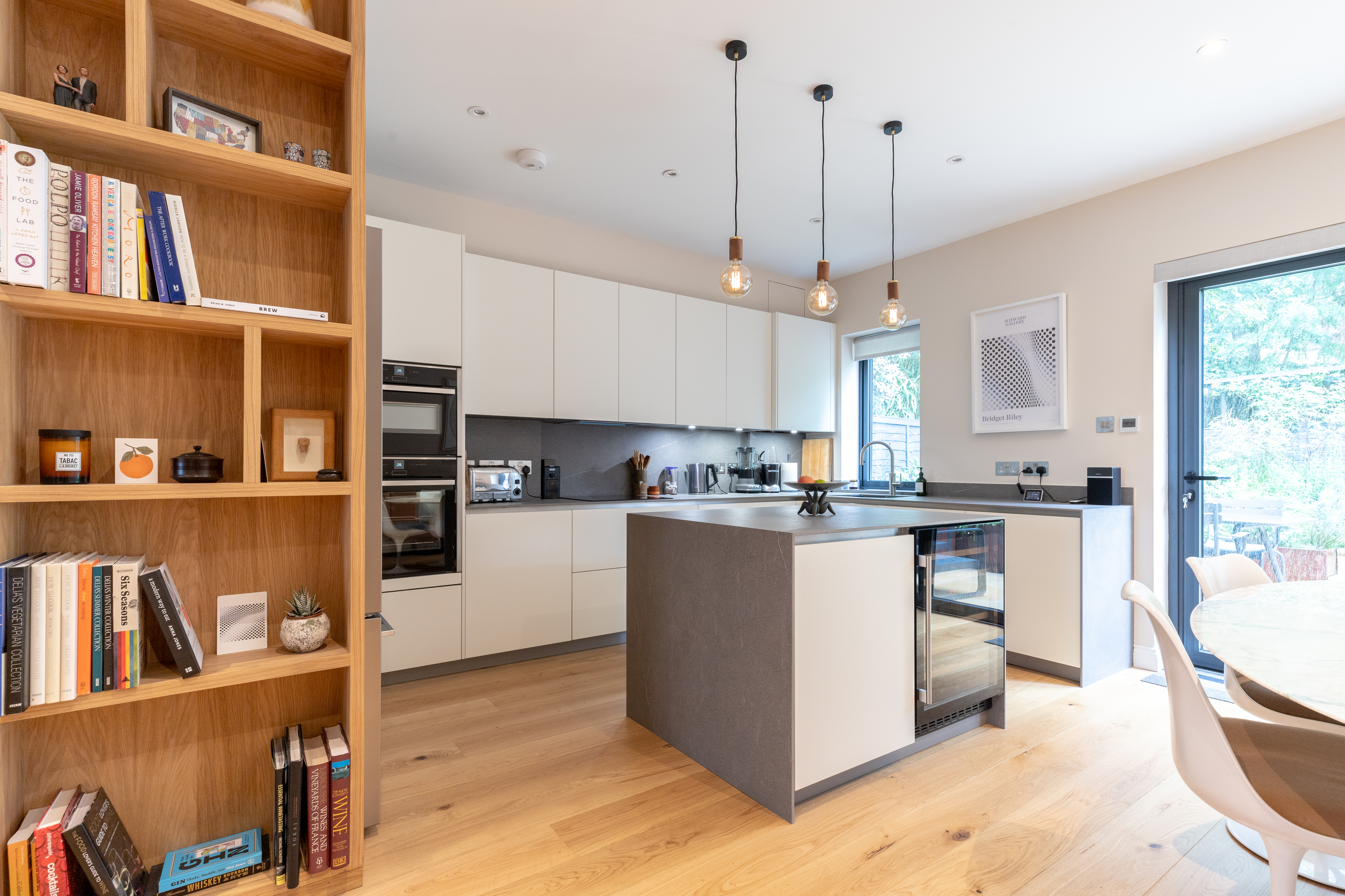 White kitchen featuring grey waterfall island worktop and upstand, Tala pendant lights, and bespoke veneer shelving to define the space