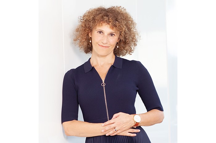 A woman with curly hair, wearing a navy blue dress and a watch, poses confidently with her hands clasped in front.