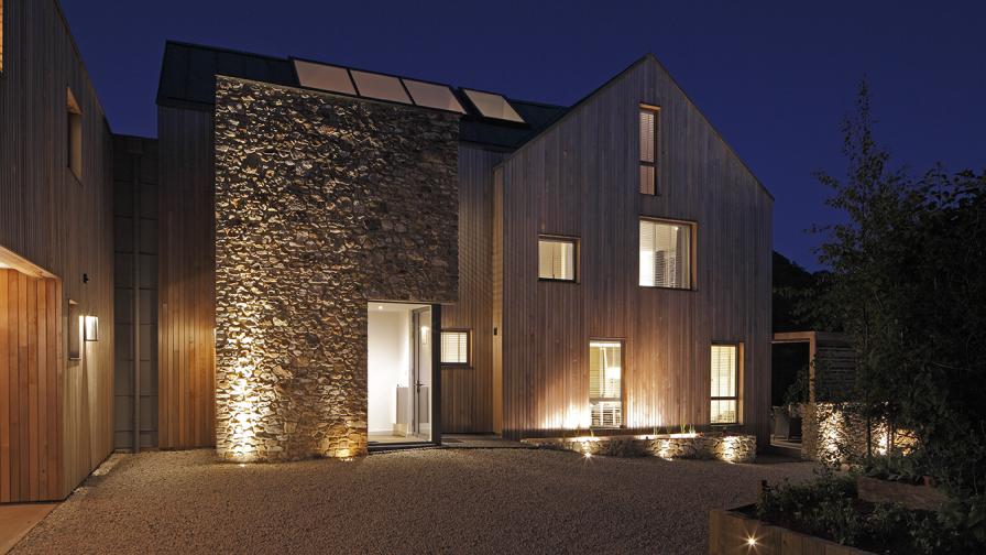 Modern house in timber and stone, at dusk with lights in the windows and exterior lighting against a dark blue sky.