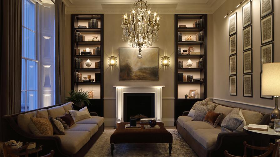 Grand regents park drawing room with two sofas either side of marble fireplace and chandelier. Recessed lit shelving along back wall. Blue dusk light at the full height sash window on the left.