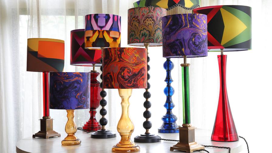 A collection of lamps with colourful patterned shades and coloured glass stands on a circular table in front of windows.