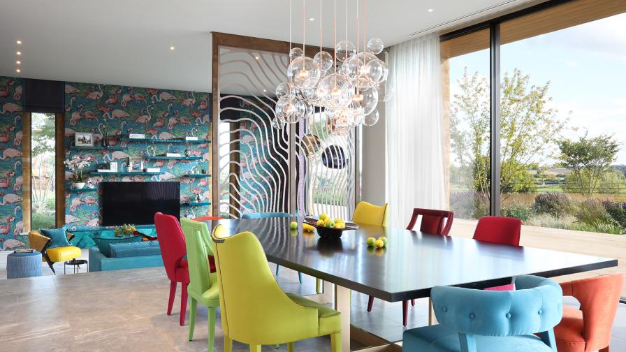 Open plan dining area with sunken seating area beyond a wavy metal screen. Mix and match coloured upholstered chairs around the table.