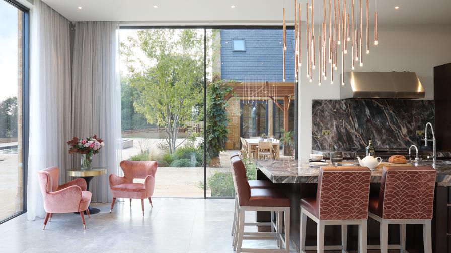 Brown marble kitchen with terracotta coloured upholstered barstools at central island. Full height sliding glass doors framed by sheer curtains frame the garden beyond.