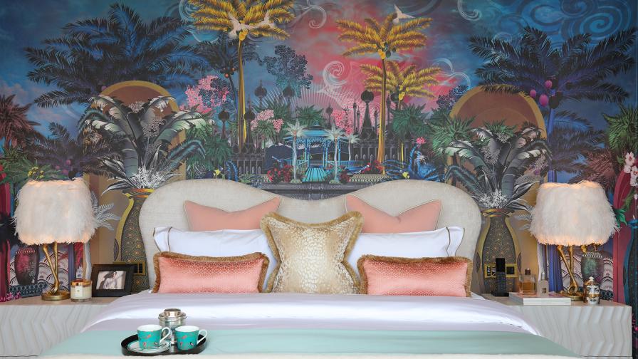 Opulent master bedroom with wallpaper fantasy panorama with palm trees and exotic architecture. A tray with coffee cups sits on the bed, and fluffy bird like lamps are on the bedside tables.