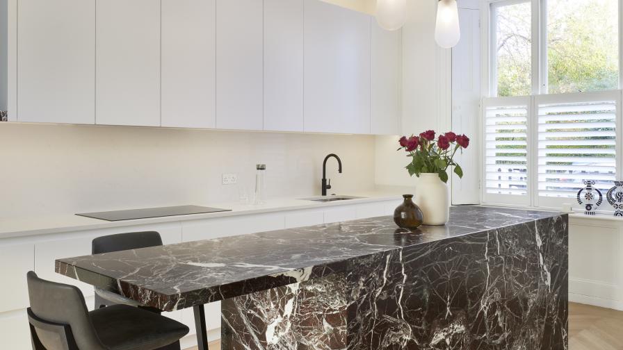 An angled view of a marble clad kitchen island with bar stools & white painted cabinets