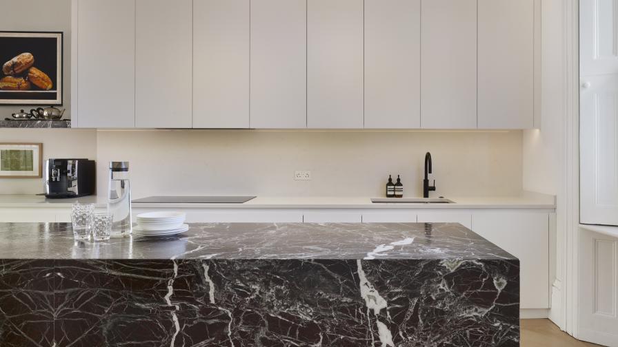 The rear view of a marble-clad kitchen island with white painted wall cabinets above the sink area against the wall in the background