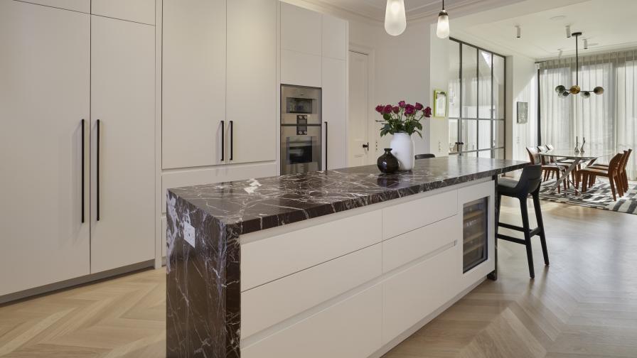 View of the front of a white painted kitchen island with a marble worktop & sides & tall cabinets on teh opposite wall.