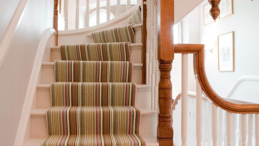 A Victorian style staircase with elaborate finials & wooden handrail with a multishade green striped carpet runner.