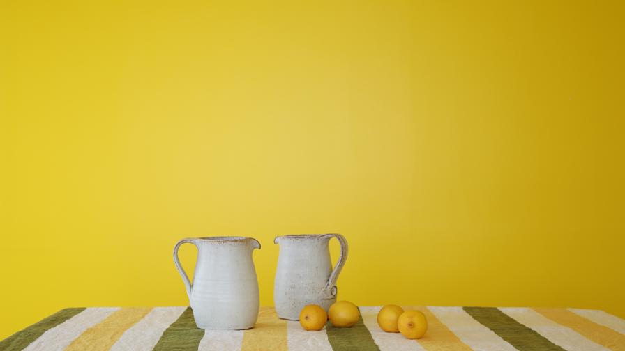 Two cream earthenware jugs and some lemons on a green and yellow striped tablecloth against a bright yellow backdrop