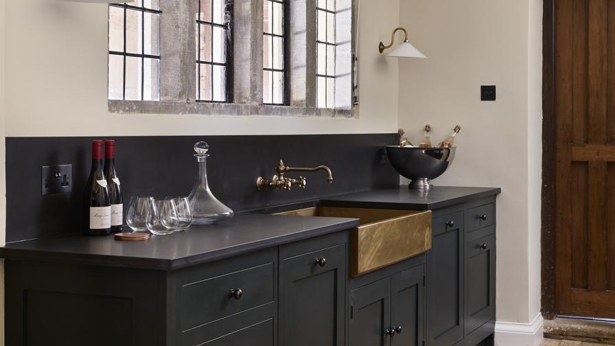 A run of dark grey traditional base cabinets with a large copper rectangular sink, decorative wall lights, & traditional red runner