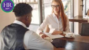 a woman in a white shirt speaks with a man in a waistcoat over a conference table