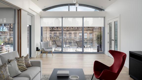 A modern living room with large floor-to-ceiling windows, white roller blinds, a gray sofa, a red accent chair, light wood flooring, and a view of a historic building outside.